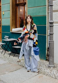 Cardigan oversized com padrão multicolorido, calças listradas em preto e branco, boné azul, acessórios incluem uma bolsa azul e várias peças de roupa.