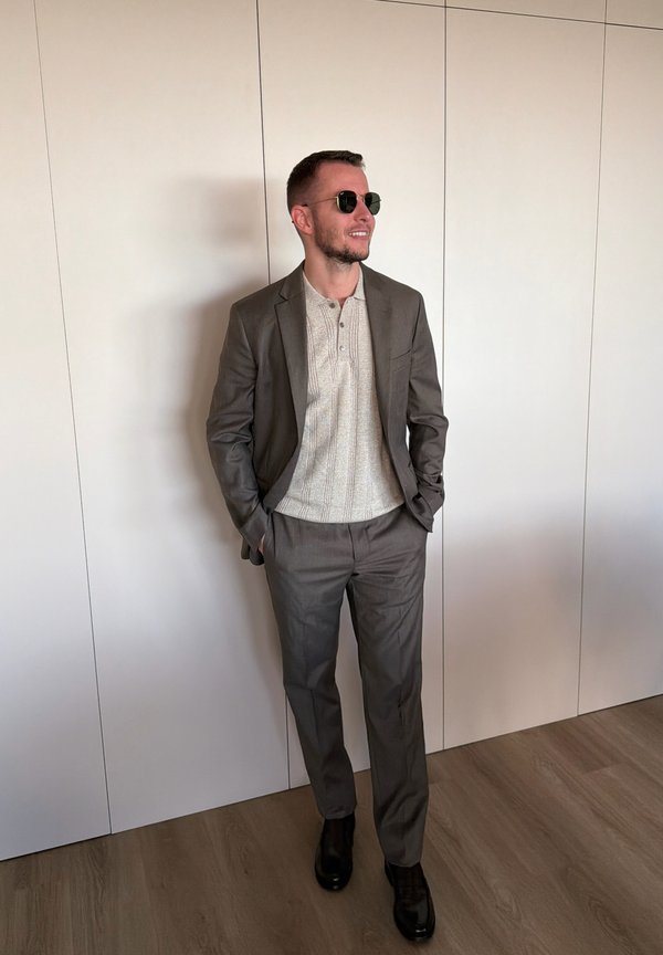 Hombre con traje gris, camisa de punto beige, zapatos negros y gafas de sol, de pie con las manos en los bolsillos frente a una pared blanca.