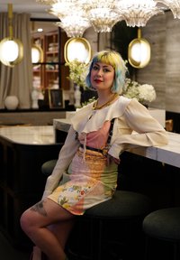 Μια γυναίκα ντυμένη με ένα πολύχρωμο φόρεμα εμπνευσμένο από λουλούδια και με ριχτή λευκή μπλούζα κάθεται σε μια πράσινη επενδεδυμένη σκαμνί σε έναν σύγχρονο εσωτερικό χώρο.