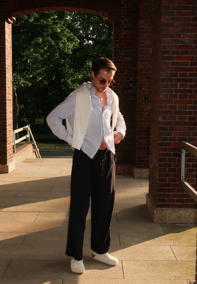 Chaleco de lino blanco sobre una camisa de color azul claro con cuello abierto, combinado con pantalones oscuros de pierna ancha y zapatos blancos. Ambiente natural al aire libre.