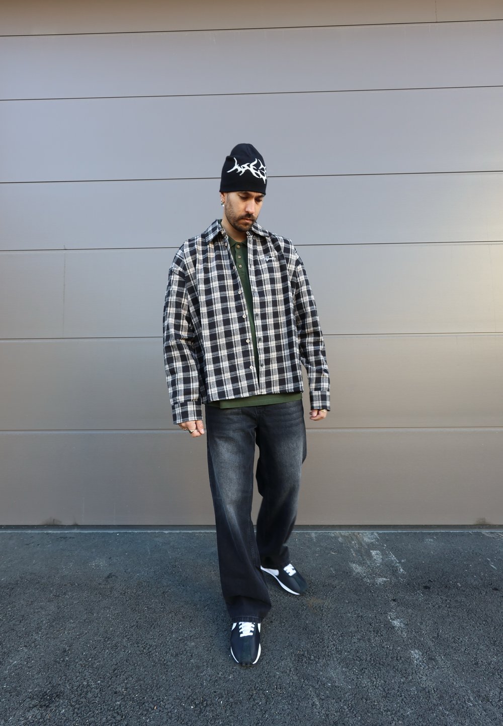 Zwart-wit geblokte oversized shirt, groene polo eronder, donkere jeans en zwarte sneakers met witte veters, gecombineerd met een zwarte beanie.