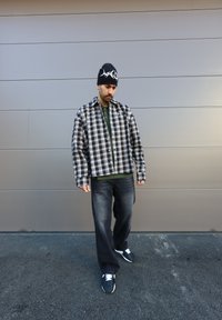 Zwart-wit geblokte oversized shirt, groene polo eronder, donkere jeans en zwarte sneakers met witte veters, gecombineerd met een zwarte beanie.