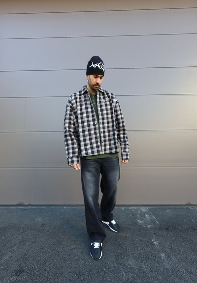 Camisa de cuadros en blanco y negro, talla oversized, polo verde debajo, jeans oscuros y zapatillas negras con cordones blancos, combinadas con una gorra de lana negra.