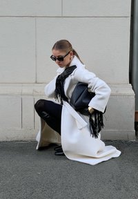 Λευκό υφασμάτινο παλτό, μαύρα δερμάτινα γάντια, μαύρη τσάντα με φούντες, μαύρα γυαλιά ηλίου και στενά μαύρα παντελόνια. Γονάτισμα μπροστά σε ανοιχτόχρωμο τοίχο.