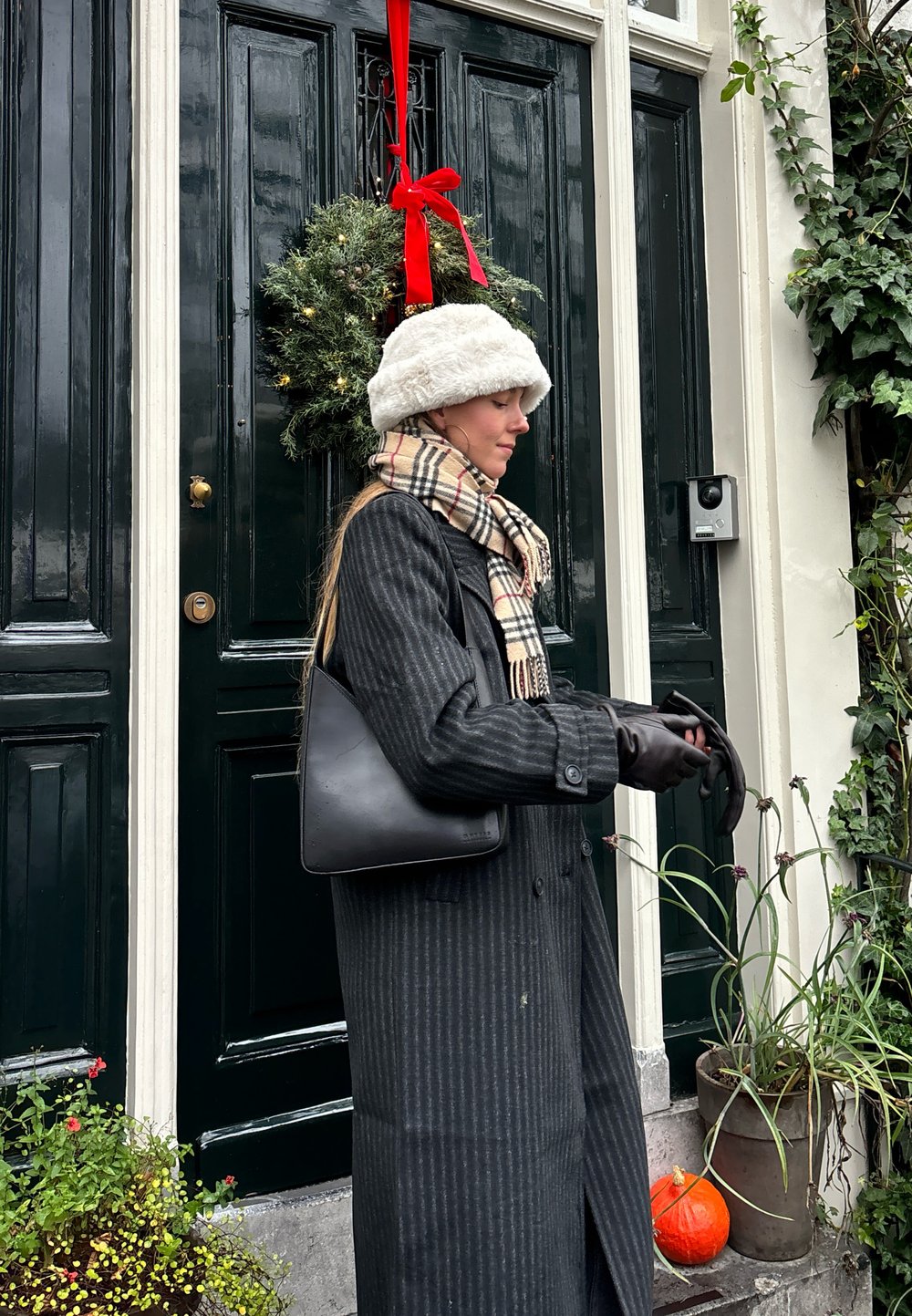 Schwarze Lederhandtasche, langer gestreifter grauer Mantel, weiße flauschige Mütze und kariertes Schal. Grüne Tür mit einem Weihnachtskranz und dekorativen Pflanzen.