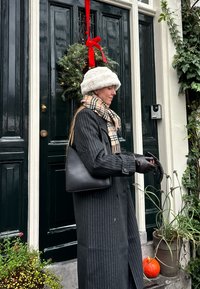 Schwarze Lederhandtasche, langer gestreifter grauer Mantel, weiße flauschige Mütze und kariertes Schal. Grüne Tür mit einem Weihnachtskranz und dekorativen Pflanzen.