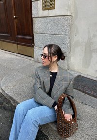 Blazer a scacchi in tonalità terrose, top nero, jeans blu, tiene una borsa marrone texture con fori circolari. Lo sfondo presenta un muro di pietra.
