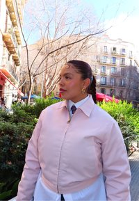 Femme aux cheveux tressés et boucles d'oreilles en or en forme de créoles, portant une veste rose clair, debout en plein air sur une place de la ville avec des arbres sans feuilles et des bâtiments en arrière-plan.
