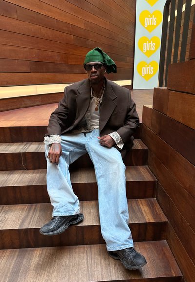 Blazer oversized marrón oscuro sobre una camisa de estampado, jeans baggy azules claros, gorro verde y zapatillas negras, sentado en escalones de madera.