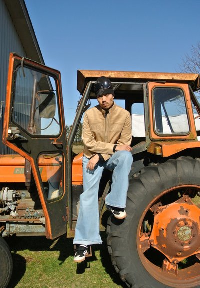 Una persona se sienta en un tractor vintage naranja, vistiendo una chaqueta cremallera color beige, jeans azul claro y zapatillas negras. El fondo es un cielo despejado.