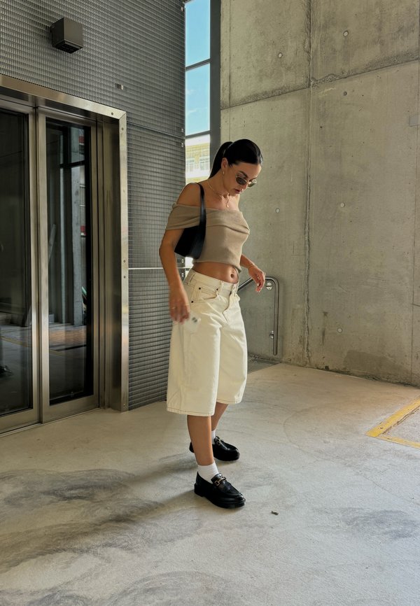 Jasnobrązowa bluzka typu off-the-shoulder, białe szorty z szerokimi nogawkami, czarne buty na poziomym obcasie z klamrami, pozująca w betonowym otoczeniu w pobliżu szklanych drzwi.