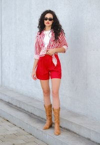 Camicetta a maniche corte a righe rosse e bianche annodata in vita sopra una maglietta bianca, abbinata a pantaloni corti rossi e stivali cowboy marroni. Tessuto liscio.