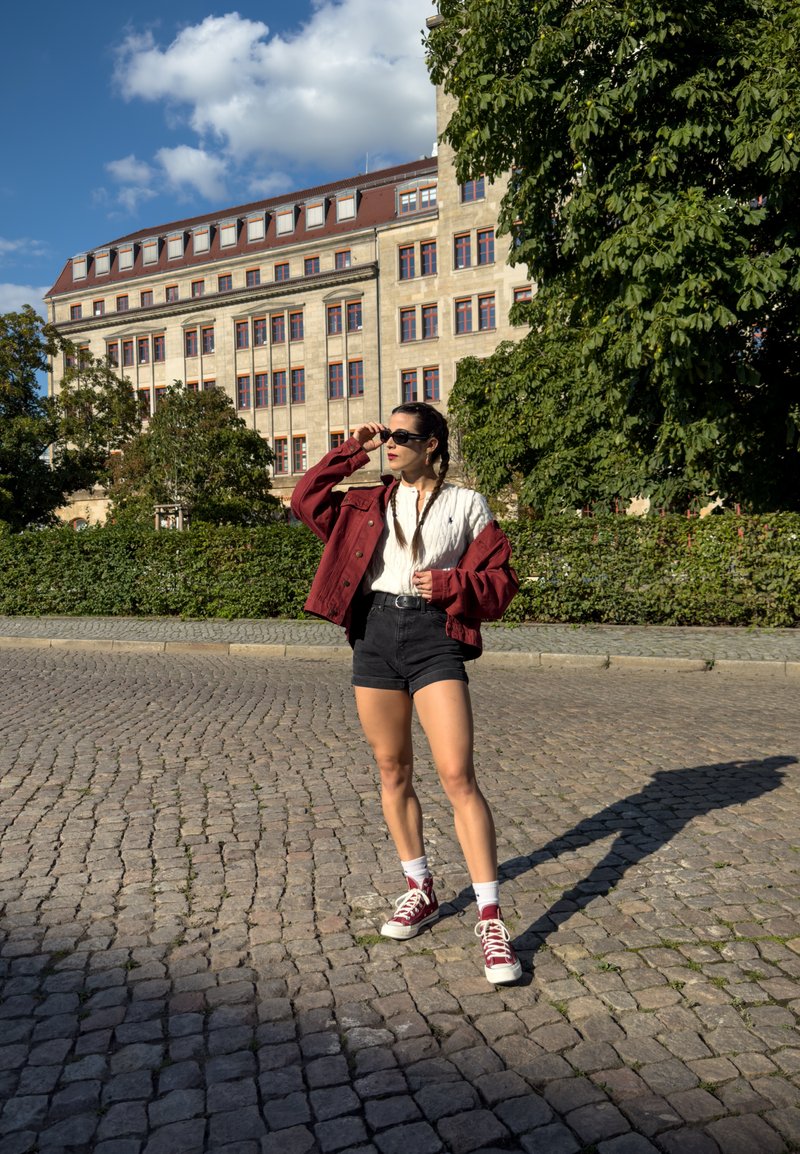 Modelo com uma jaqueta oversized em bordeaux sobre uma blusa branca, calções pretos e sapatilhas vermelhas. Chão de paralelepípedos e um edifício histórico ao fundo.
