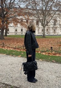 Črna usnjena jakna s kratkim krojem, kombinirana s črnimi hlačami in teksturirano črno torbico. V ozadju jesensko listje.