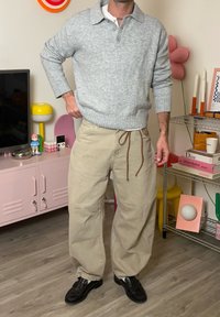 Hall halli kampsun kraega, beežid paelaga püksid ja mustad kingad. Minimalistlik tekstuur ja lihtne sobivus, paikneb värvikirevas ruumis.