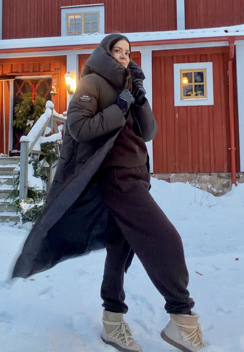 Lang sort dunjakke med høj krave, båret over en mørkebrun påklædning. Personen har handsker på og beige vinterstøvler, og står i sneen.