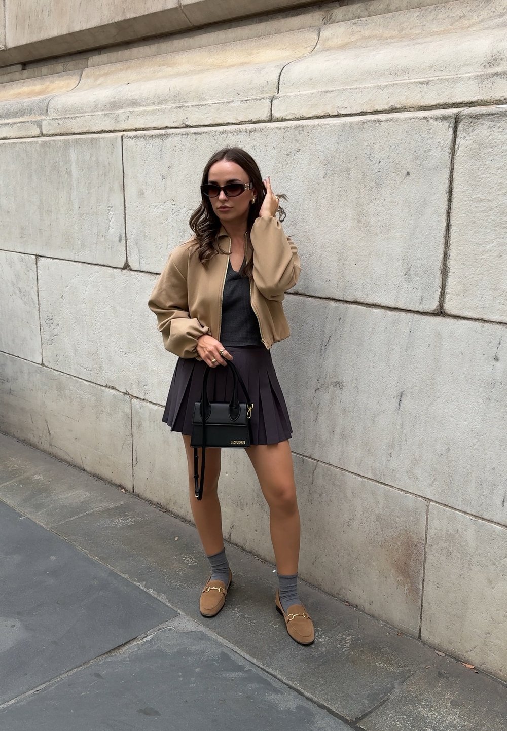 Chándal beige, camiseta gris, falda negra plisada, mocasines marrón claro y un pequeño bolso negro con detalles en dorado. Texturas suaves.