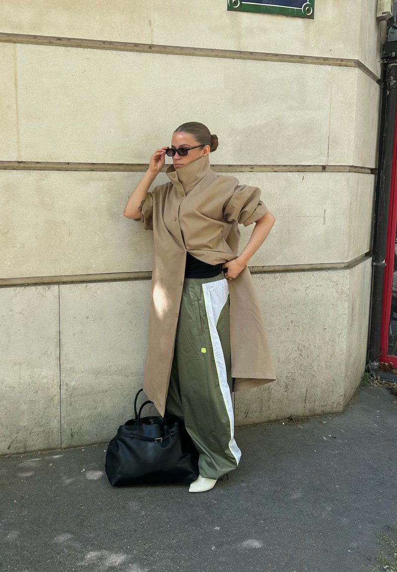 Femme appuyée contre un mur beige portant un trench-coat beige, des lunettes de soleil, un pantalon large vert et blanc, des chaussures blanches, avec un grand sac noir au sol.