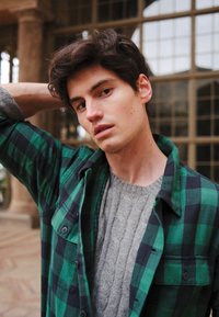 Chemise à carreaux verte et noire portée par-dessus un pull gris à torsades. La chemise est boutonnée et présente une coupe décontractée, avec une poche visible.