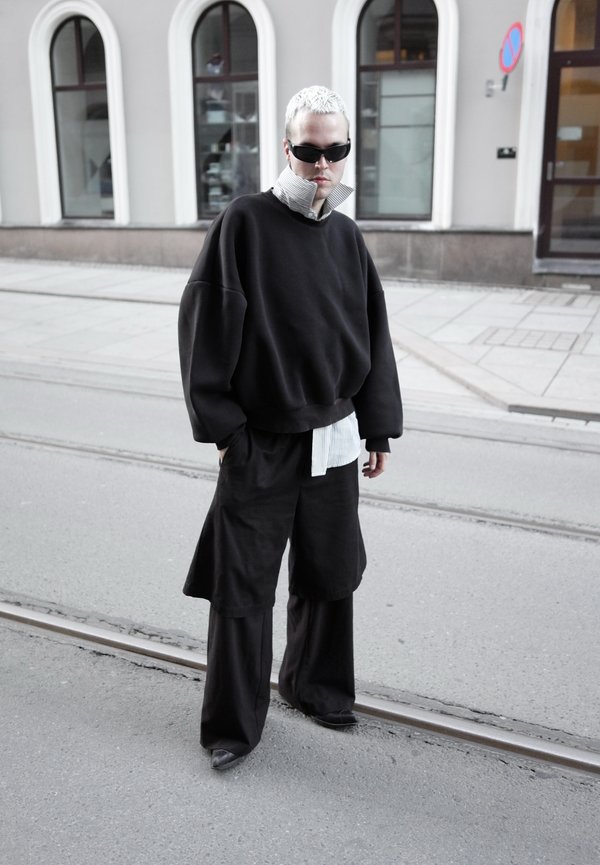Personne aux cheveux blancs courts et lunettes de soleil foncées portant des vêtements noirs superposés et surdimensionnés, debout dans une rue avec des rails de tram.