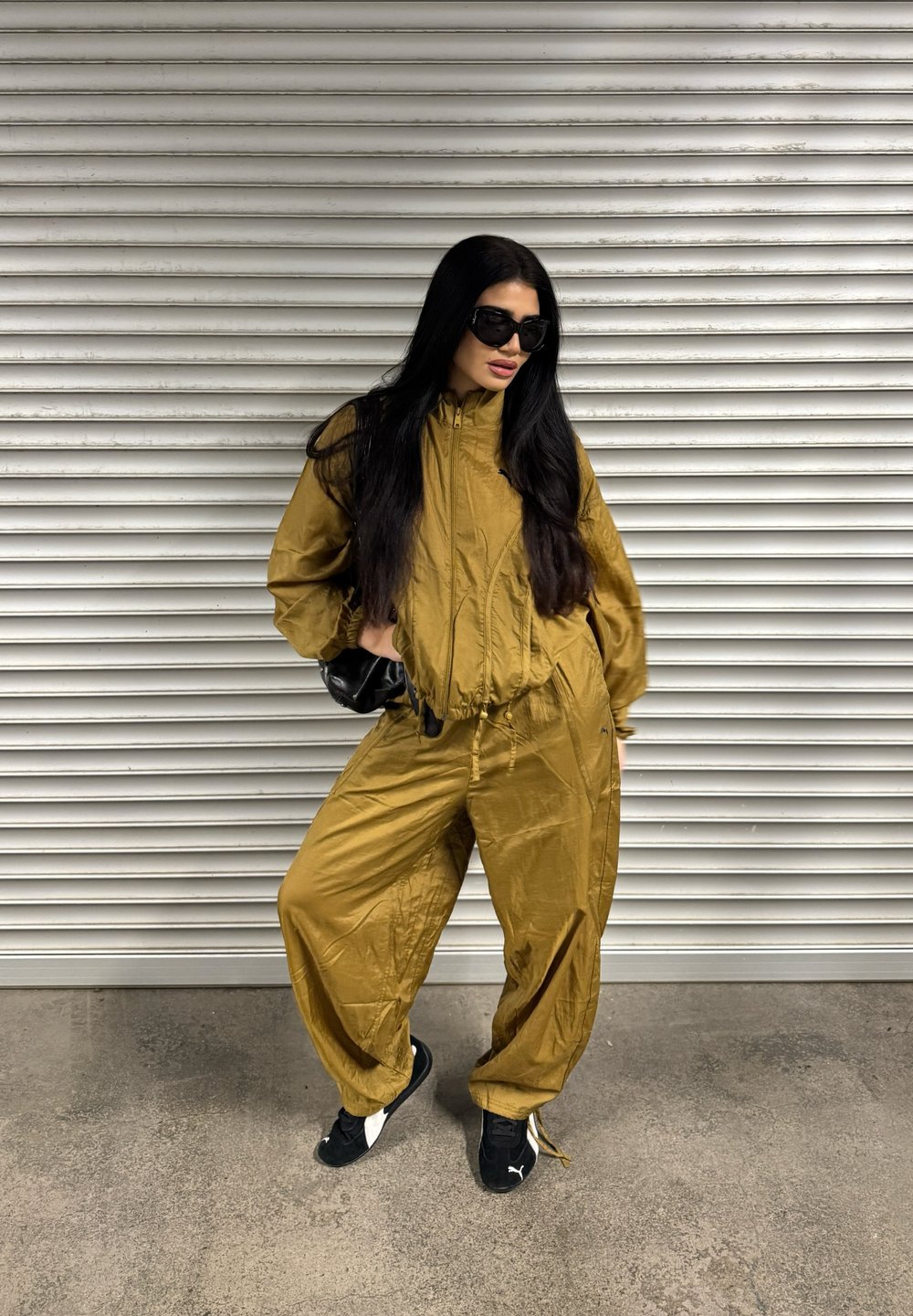 Złoty dres z zamkiem błyskawicznym i elastycznym pasem. Luźne spodnie oraz czarne sneakersy z białymi akcentami. Na twarzy czarne okulary przeciwsłoneczne.