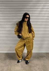 Złoty dres z zamkiem błyskawicznym i elastycznym pasem. Luźne spodnie oraz czarne sneakersy z białymi akcentami. Na twarzy czarne okulary przeciwsłoneczne.