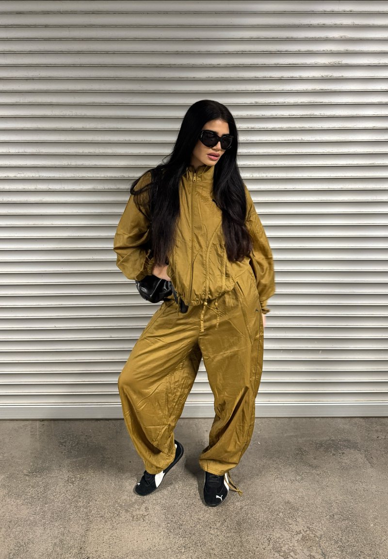 Survêtement de couleur or avec une veste zippée et une taille élastique. Pantalons amples et baskets noires avec accents blancs. Lunettes de soleil noires portées.
