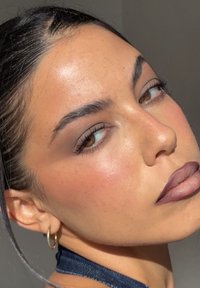 Primer plano de un rostro con piel suave y un brillo sutil. Presenta labios marrones mate y audaces, cejas definidas y pestañas suaves. Se observa un pendiente de aro dorado.