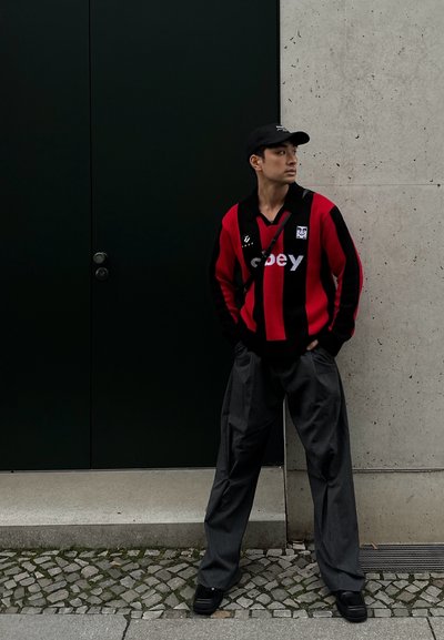 Suéter a rayas rojas y negras con un cuello negro, combinado con pantalones holgados de rayas gris claro y deportivas negras. Sombrero llevado ligeramente de lado.