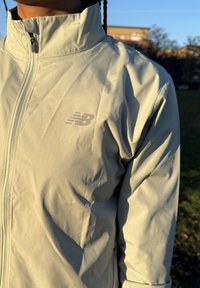 Veste de sport gris clair avec un col montant, fabriquée en matériau lisse et léger. Elle présente un logo discret sur la poitrine et une fermeture éclair.