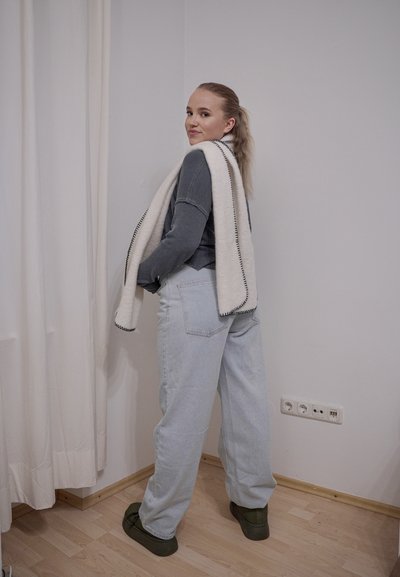 Vaqueros extragrandes de color gris claro combinados con un top de manga larga acanalado de color gris oscuro y una bufanda blanca con ribete negro, sobre un fondo neutro.