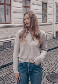 Lys grå cardigan med glidelås, ribbet tekstur og sidelommer, kombinert med blå jeans. Mykt strikket stoff, avslappet passform og lange ermer.