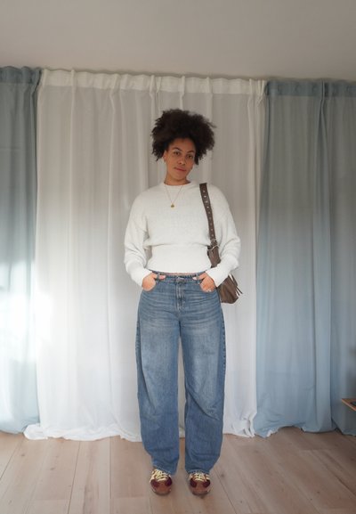 Suéter blanco de punto con dobladillo acanalado, vaqueros anchos de tiro alto en azul, bolso marrón de hombro y zapatillas marrones con cordones amarillos.