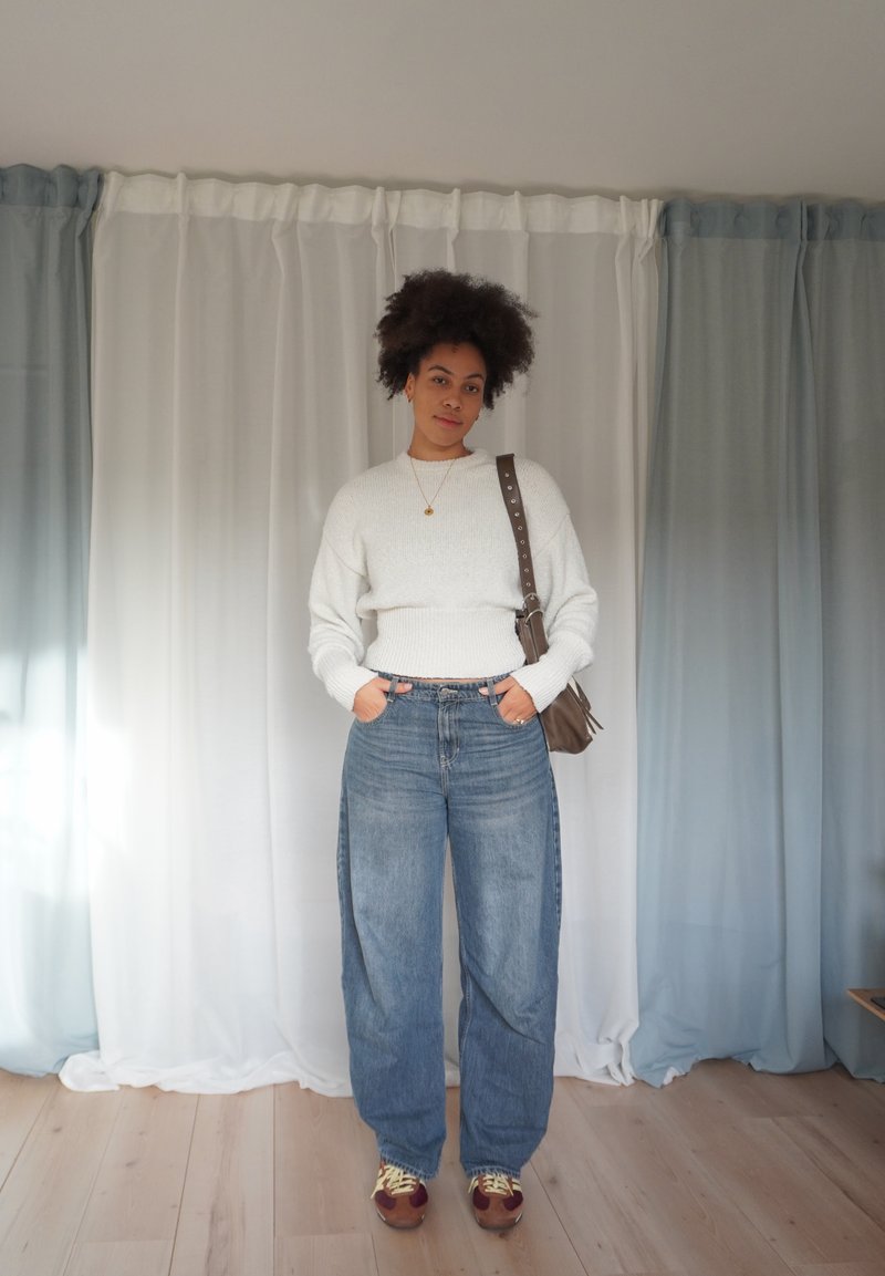 Maglione bianco a maglia con bordo a coste, jeans blu a gamba larga a vita alta, borsa marrone a tracolla e sneakers marroni con lacci gialli.