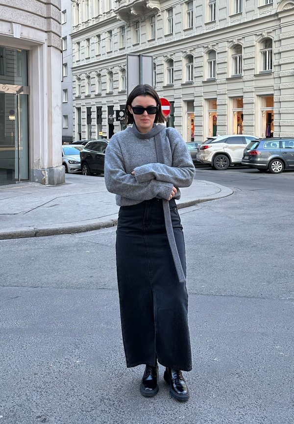 Šedý pletený svetr s vysokým límcem, dlouhá tmavá denimová sukně a černé šněrovací boty, stojící na městské ulici.