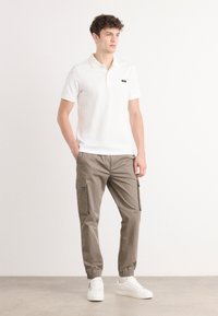 Bela polo majica s kratkimi rokavi in majhnim črnim logotipom, v kombinaciji s khaki cargo hlačami, ki imajo stranske žepke in elastične manšete, obute z belimi športnimi copati.
