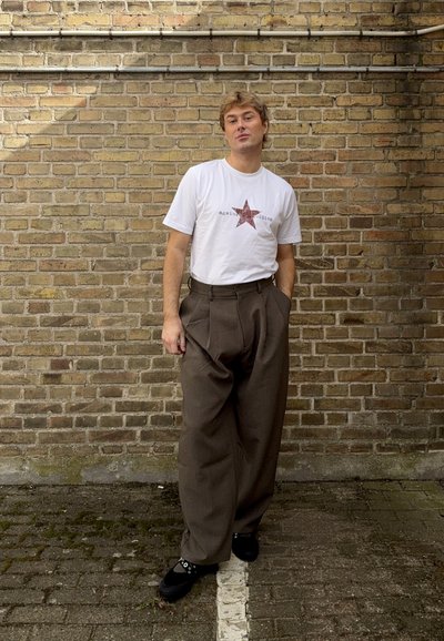 Joven de pie al aire libre contra una pared de ladrillos, vistiendo una camiseta blanca con una estrella roja y pantalones marrones de pierna ancha.