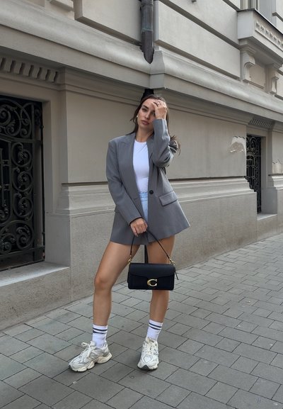 Blazer gris ajustado sobre una camisa blanca, combinado con pantalones cortos a rayas, zapatillas blancas y un bolso de hombro negro con detalles dorados. Fondo de hormigón.