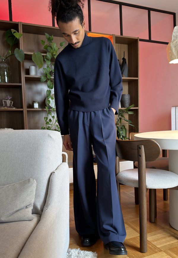Homme aux cheveux bouclés attachés en chignon, portant un col roulé bleu marine et un pantalon large, debout dans un salon moderne avec des plantes et des meubles en bois.