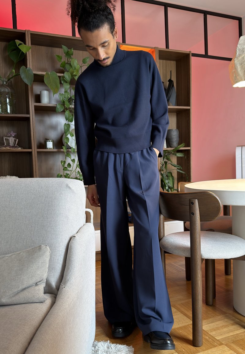 Kiharatukkainen mies nutturalla, pukeutunut tummansiniseen poolopaitaan ja leveälahkeisiin housuihin, seisoo modernissa olohuoneessa, jossa on kasveja ja puisia huonekaluja.