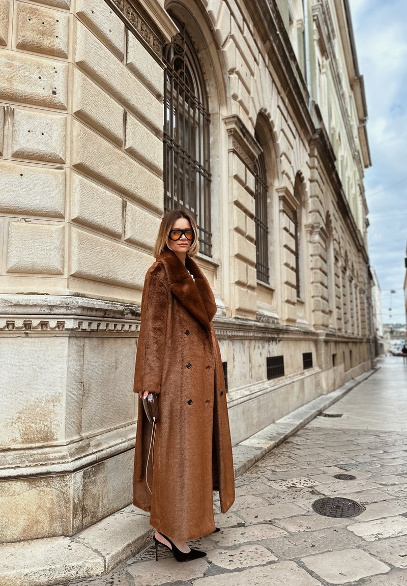 Brauner, zweireihiger Mantel mit großem Kragen aus Plüschstoff. Getragen über einem langen Kleid, kombiniert mit schwarzen Pumps und einer metallischen Clutch.