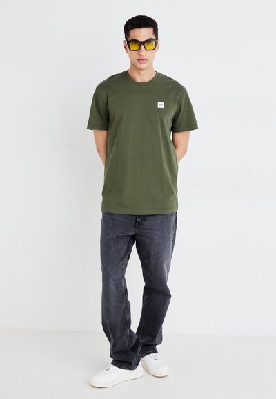 Camiseta de manga corta verde oliva con un pequeño logo en el pecho, combinada con jeans gris oscuro y zapatillas blancas; el modelo lleva gafas de sol amarillas.