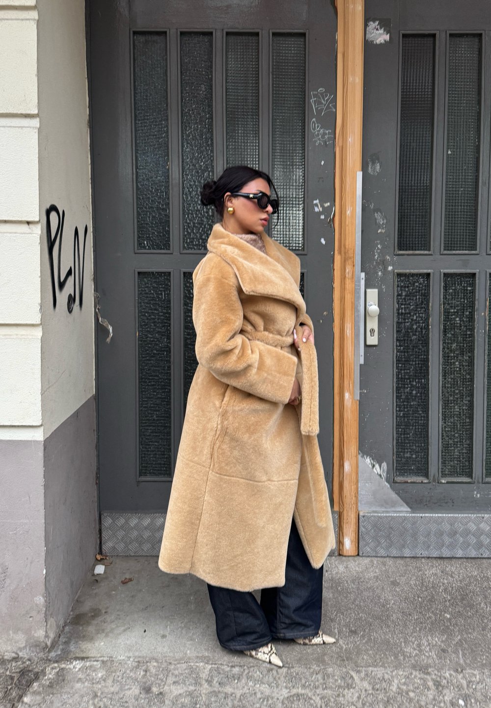 Μπεζ ψεύτικο γούνινο παλτό με γιακά σάλι και ζώνη, σε συνδυασμό με φαρδιά σκούρα μπλε τζιν και σχέδιο παπούτσια. Φόντο γκρίζας πόρτας.