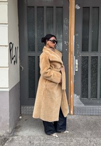 Μπεζ ψεύτικο γούνινο παλτό με γιακά σάλι και ζώνη, σε συνδυασμό με φαρδιά σκούρα μπλε τζιν και σχέδιο παπούτσια. Φόντο γκρίζας πόρτας.