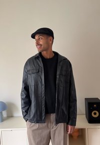 Fekete bőr ingdzseki két mellzsebbel, fekete pulóver fölött viselve. Sötét nadrág és fekete lapos sapka egészíti ki a megjelenést.