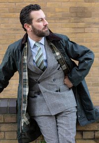 Grey plaid double-breasted suit layered beneath a dark green waxed jacket with a brown collar; light blue shirt and striped tie, leaning casually.