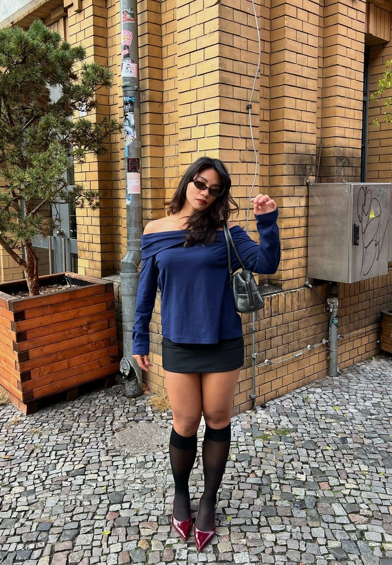 Top blu navy scoperto sulle spalle, mini gonna nera, calze sheer fino alla coscia e décolleté bordeaux. Borsa scura, sfondo di muro in mattoni testurizzati.