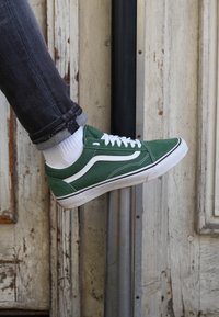 Sneaker in tela verde con dettagli e lacci bianchi, suola in gomma, design a profilo basso, abbinato a jeans grigi e calzini bianchi.