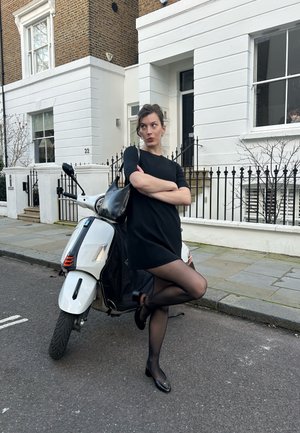 Mujer con vestido negro y medias cruzando los brazos, apoyada en un scooter blanco estacionado en la calle frente a una casa adosada blanca con barandilla negra.