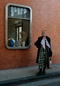 Osoba w czarnym płaszczu i wzorzystej sukience stoi przy dużym oknie z metalową ramą, na tle pomarańczowej, o fakturze ściany.